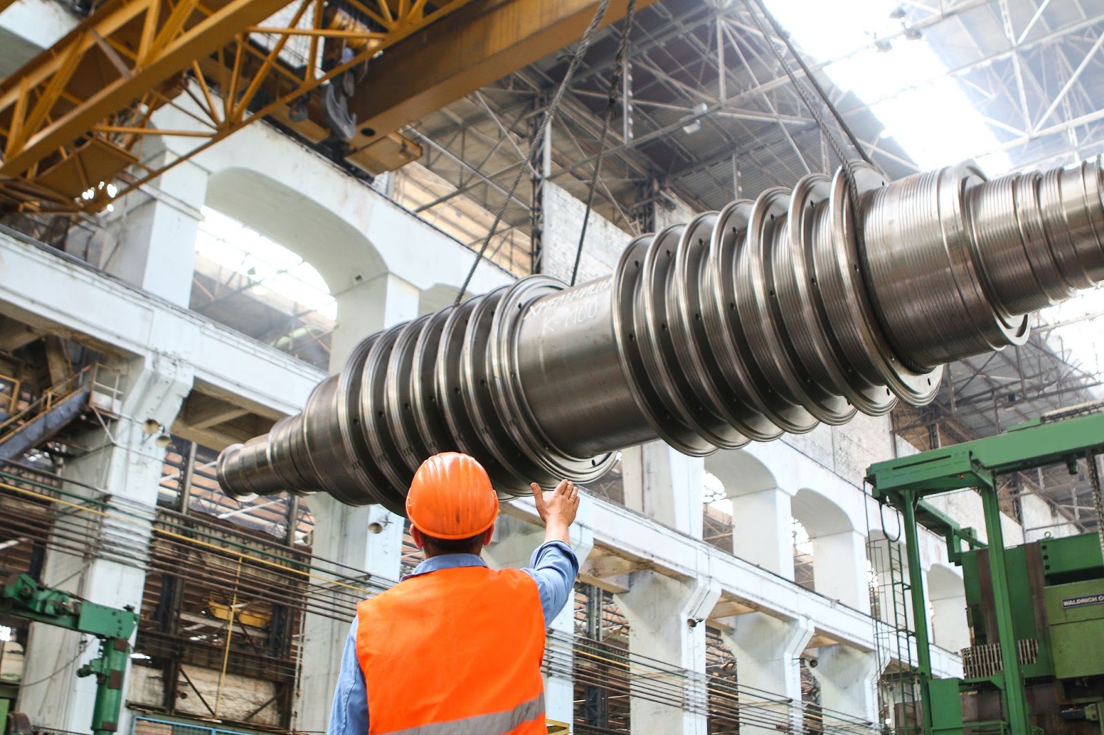 Man stands under a large piece of equipment at a manufacturing plant while wearing protective gear.